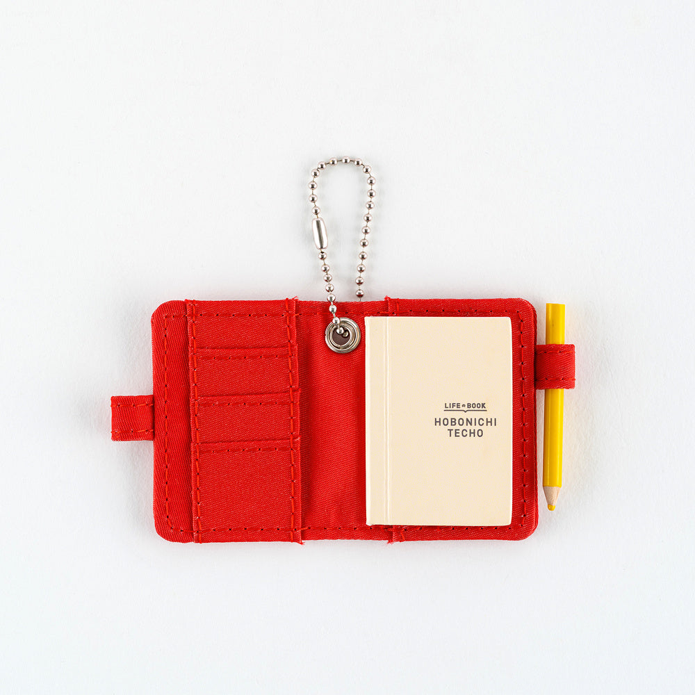 Open small red notebook keychain with a silver ball chain and a red loop holding a yellow pencil, with mini Hobonichi Techo planner inside shown on a plain white background.