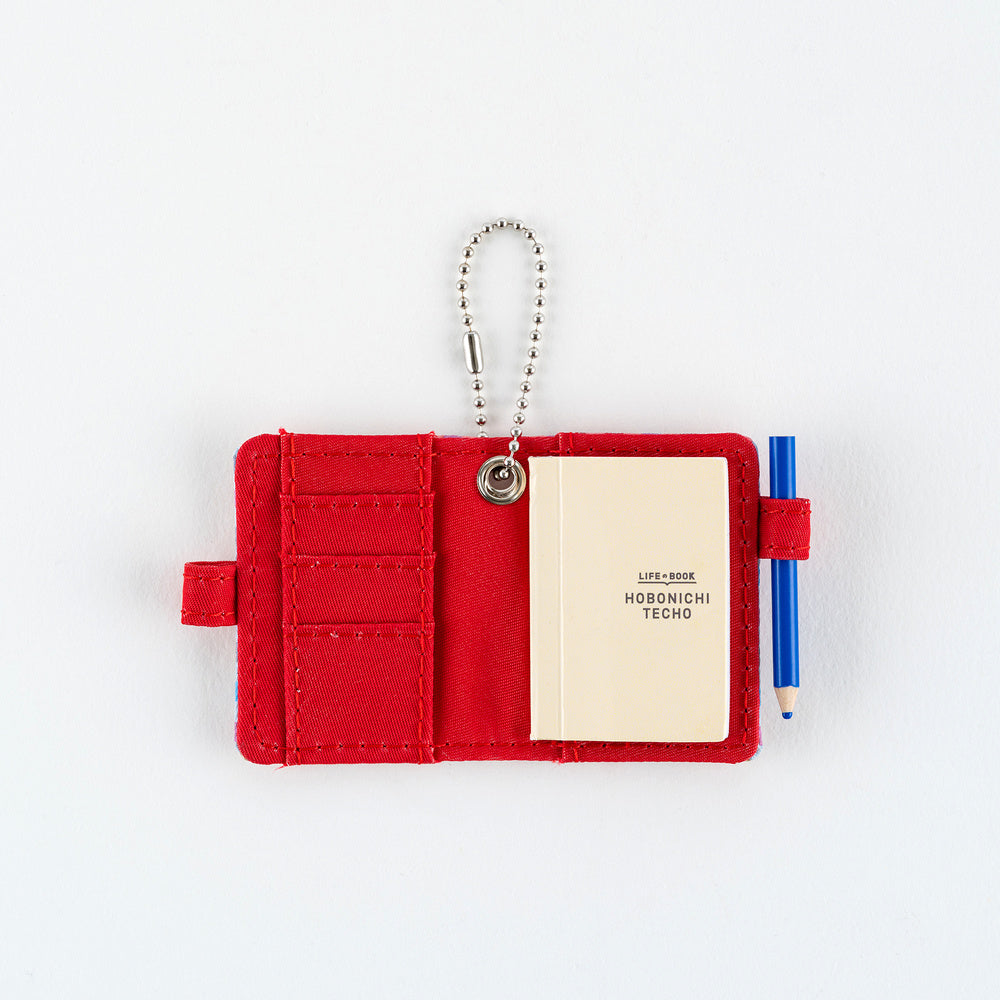 Open small red notebook keychain with a silver ball chain and a red loop holding a blue pencil, with mini Hobonichi Techo planner inside shown on a plain white background.