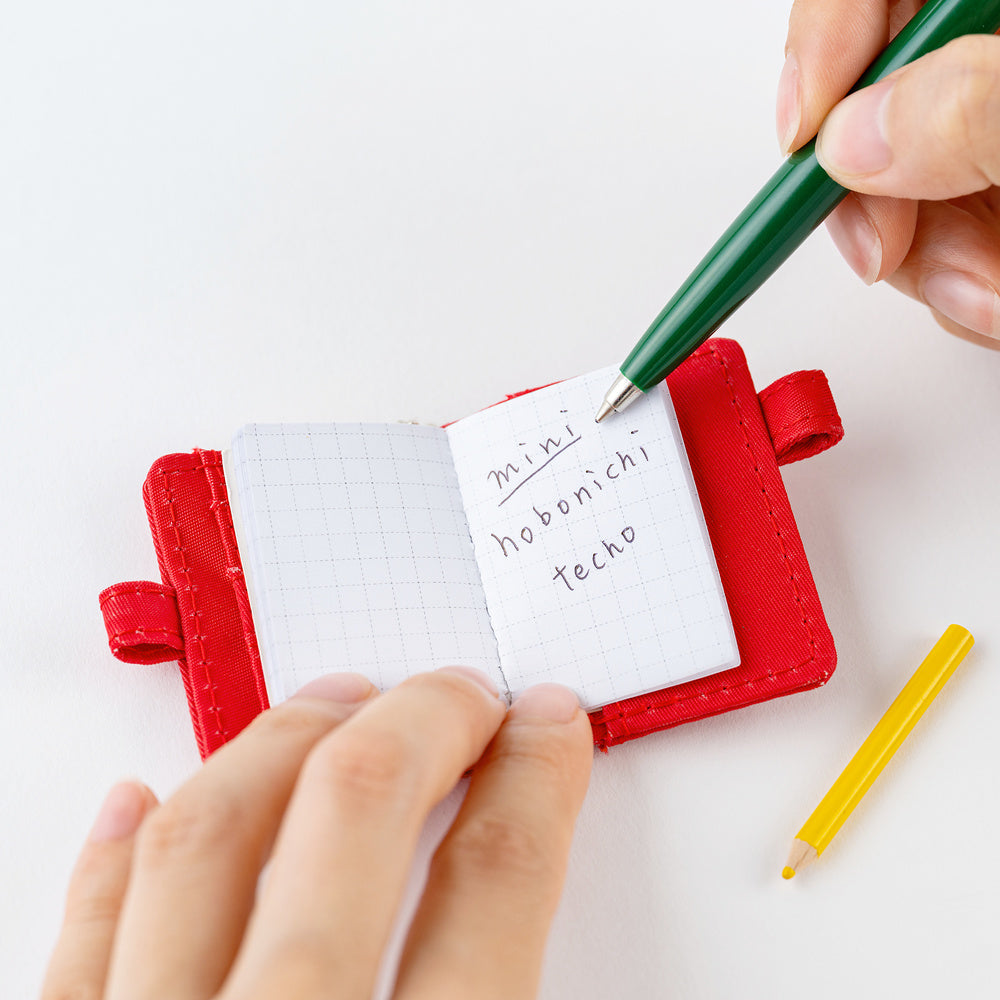 A miniature red notebook with a grid page, held open by hands, with a green pen writing on the cover and a yellow pencil on a white background.