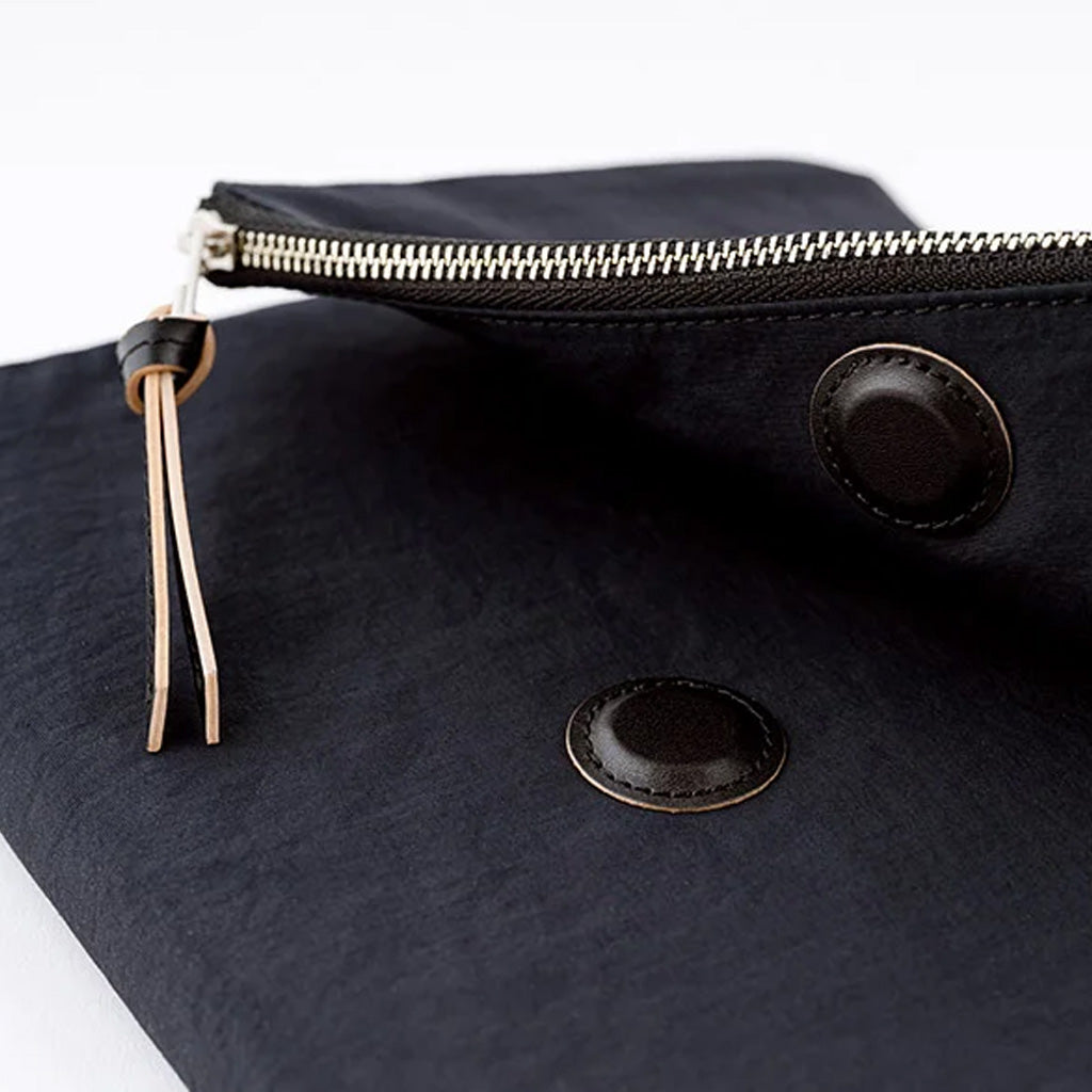 Close-up of a black fabric pouch with a silver zipper, a light tan leather pull tab, and two round black snap buttons on a white background.