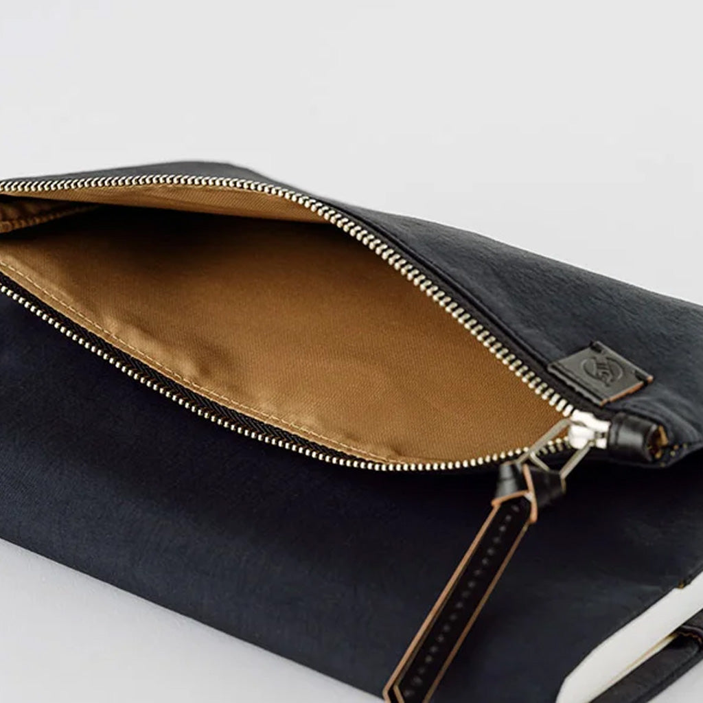 A close-up of a black zippered pouch shown open, revealing a tan interior lining and a gold-toned zipper. A small leather tag with a monogram logo is attached near the zipper pull.
