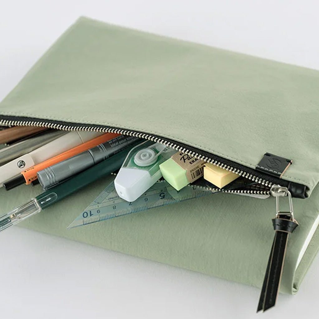 A sage green canvas zippered case shown open, filled with stationery items including pens, a correction tape dispenser, a ruler, and sticky notes, on a clean white background.