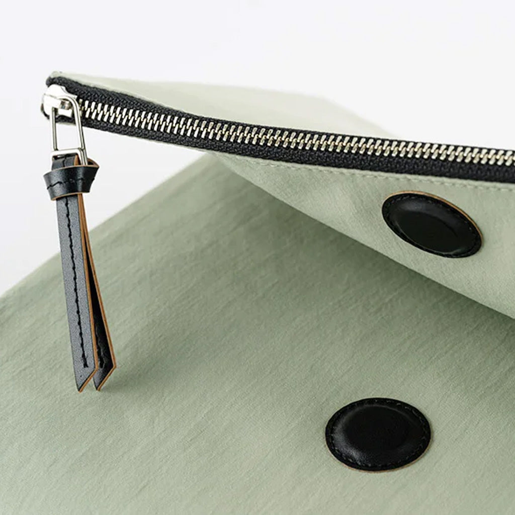 A close-up of a sage green fabric pouch with a black zipper, a black leather tassel zipper pull, and two black snap buttons.