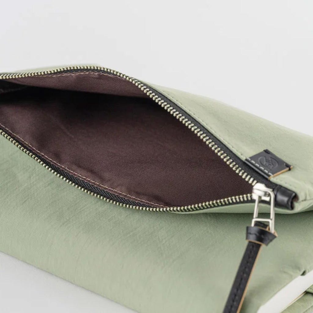 A close-up of a sage green pouch shown open, revealing a dark brown satin-lined interior with a gold-tone zipper and a small black leather brand tab.