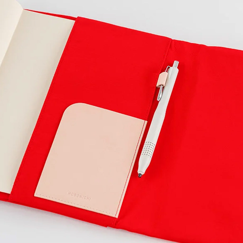A red fabric notebook cover shown open on a white background, with a cream-colored interior page, a pale blush pocket, and a white pen tucked inside.