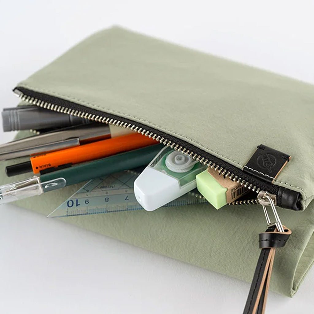 An olive green fabric zippered pouch with a black zipper and a small black brand tag, shown open with pens, a correction tape dispenser, a ruler, and a eraser inside on a white background.