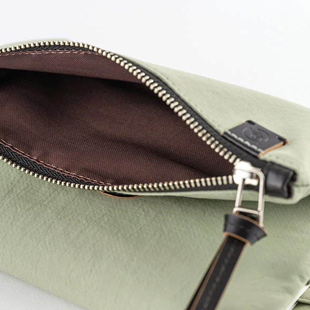 Close-up of a sage green leather pouch with an open zipper revealing a dark brown interior, featuring a silver-tone metal zipper pull and a small black leather brand tab.