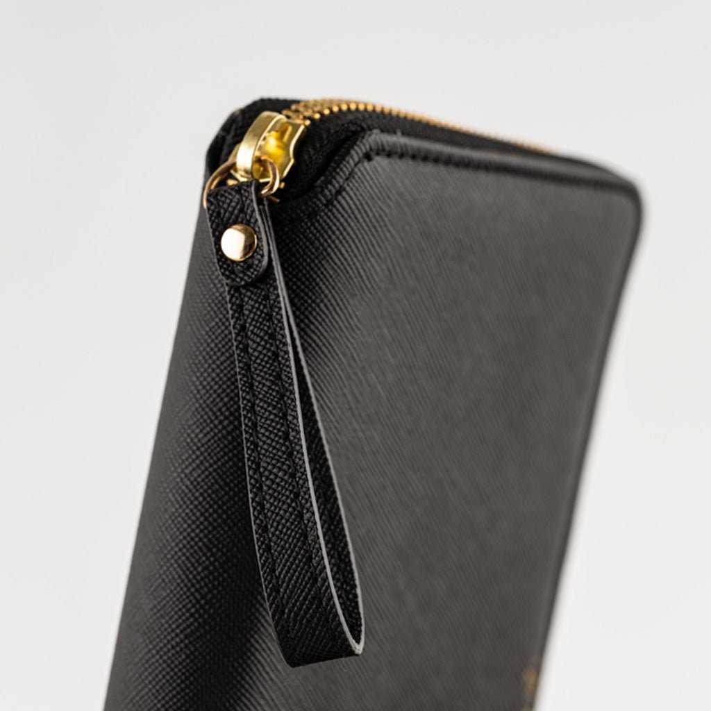 Close-up of a black textured zip-around notebook cover with a gold-tone zipper pull and wrist strap on a light gray background.