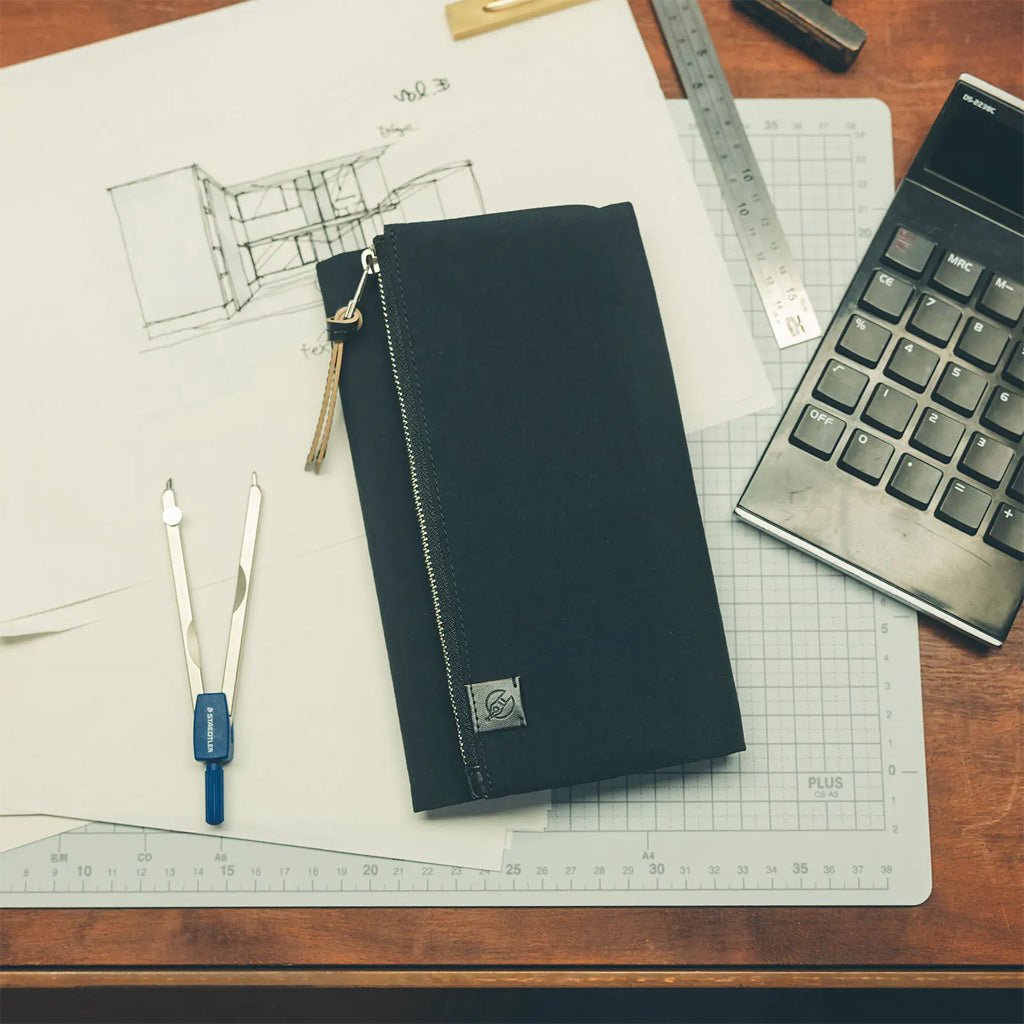 A black zippered fabric notebook cover with a small logo patch, placed on a desk with drafting tools, a calculator, and a grid cutting mat.