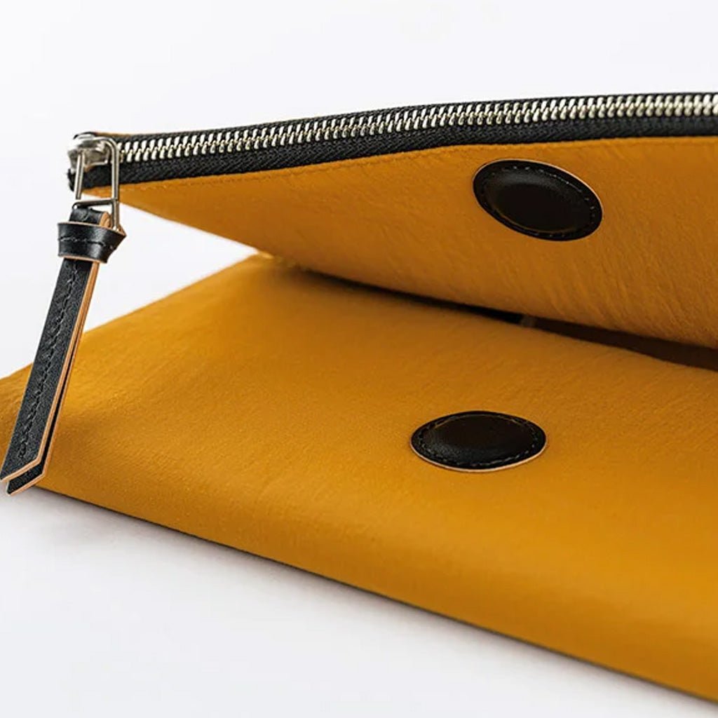 A close-up of a mustard yellow leather notebook cover with a black zipper, black circular snap details, and a black leather zipper pull on a white background.