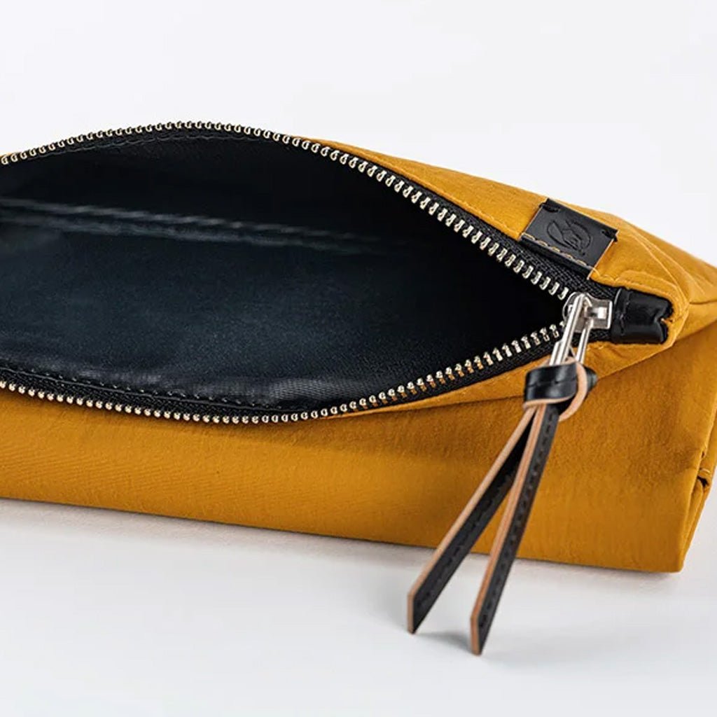 A close-up of a mustard yellow pouch shown open, revealing a black interior lining with a silver-tone zipper and a black leather pull tab on a white background.