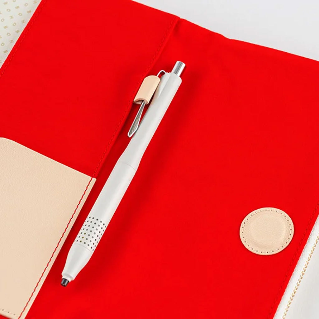 A white pen with a silver clip rests diagonally on a bright red fabric notebook cover with beige faux-leather accents.