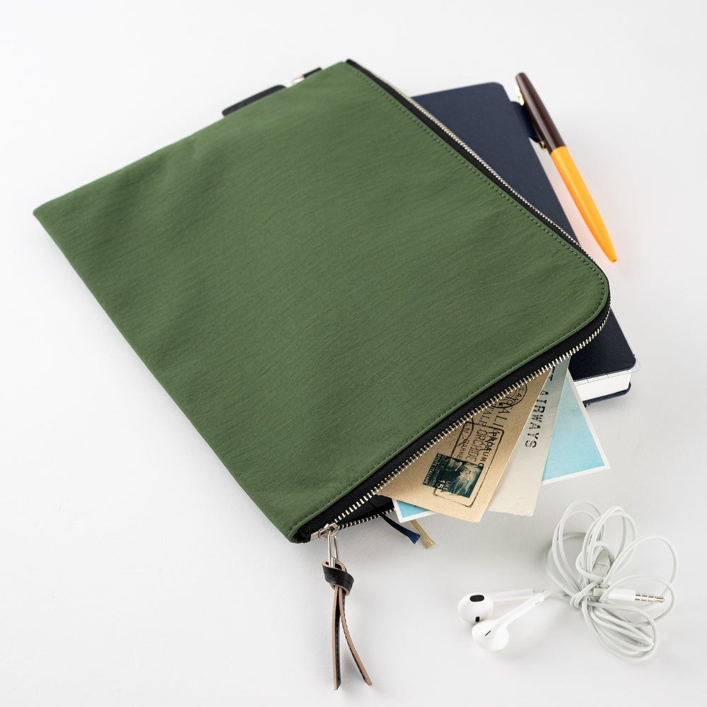 An olive green canvas laptop sleeve with a black zipper, shown open and styled with a notebook, pen, postcard, and white wired earphones on a clean white background.