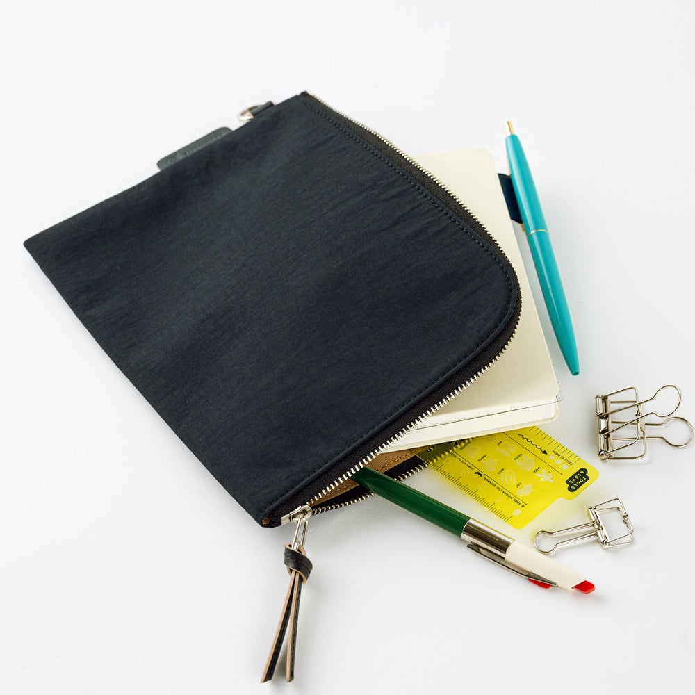 A black zippered fabric pouch with a silver zipper and tassel, shown open with stationery items like a notebook, pen, ruler, and binder clips on a white background.