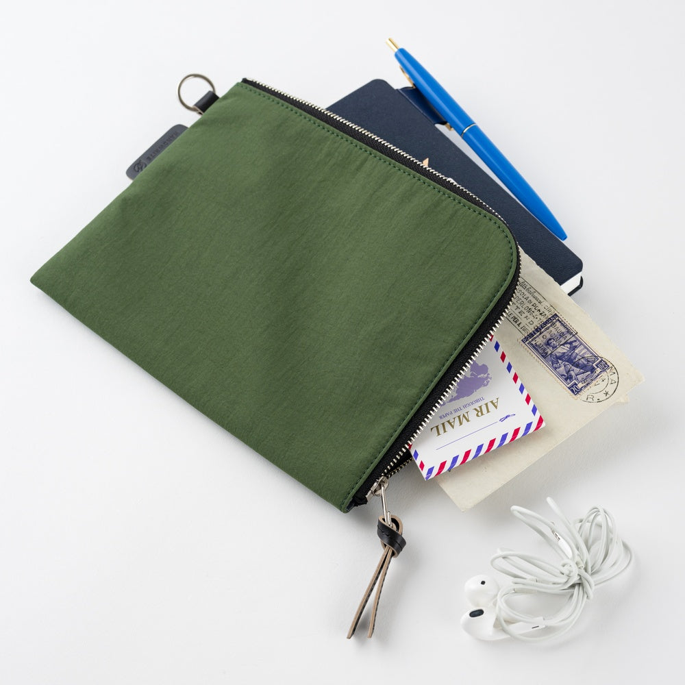 An olive green zippered pouch shown on a white background, filled with travel items including a notebook, pen, passport, and mailer, with white wired earbuds placed beside it.