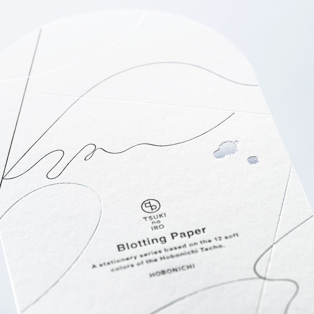 A close-up of a sheet of TSUKI no IRO Blotting Paper from the Hobonichi Techo series, featuring fine black line art, subtle gray abstract shapes, and two small blue-gray blotches on a white textured paper background.