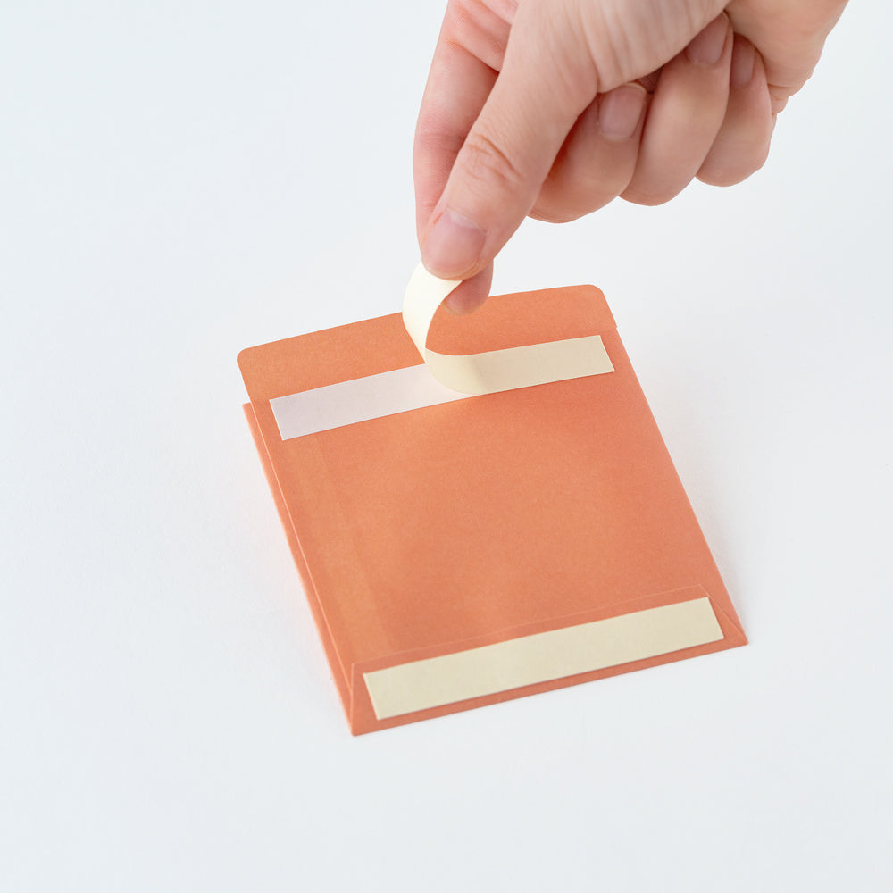 A hand peeling off a beige adhesive strip from an orange square card on a clean white background.