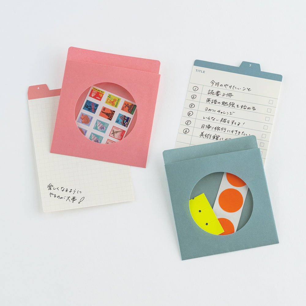 Three colorful paper envelope-style stationery pieces arranged on a white background: a pink envelope with a circular cutout showing a sheet of small illustrated stickers, a white grid notepad with handwritten text, and a muted blue envelope with a circular cutout showing a sheet of orange and yellow dots.