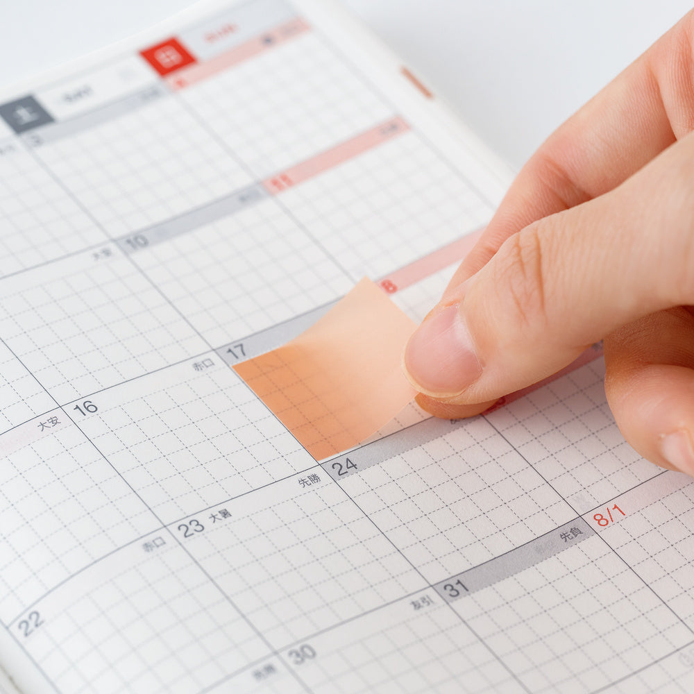A close-up of a hand applying a peach-colored adhesive sticky note to a white grid calendar planner.