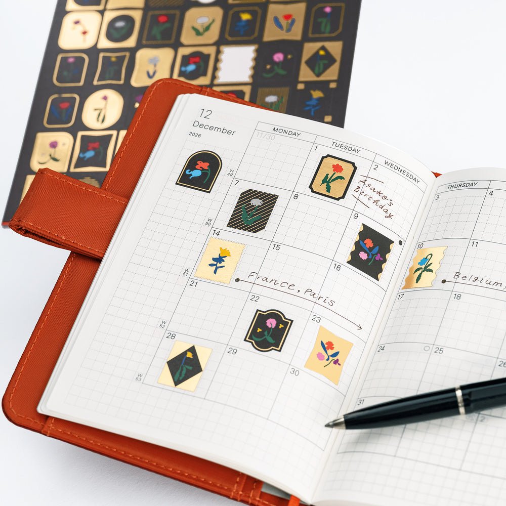 An open orange-brown planner with a grid calendar page, decorated with Yoshie Watanabe stickers with gold foiling and colorful flower motifs. A black pen rests on the page, and a sheet of matching decorative stickers is visible in the background on a white surface.