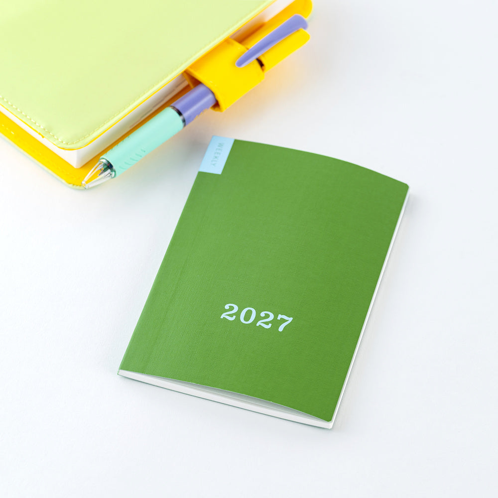 A green 2027 weekly planner with blue text on the cover, styled on a white background next to a yellow notebook cover with a pen holder with a pen.