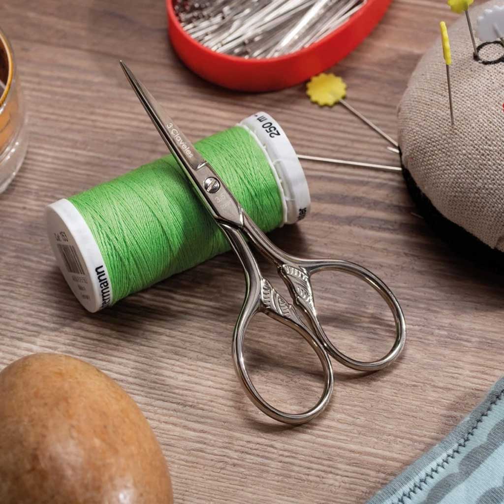 Green spool of thread with scissors on a wooden surface, surrounded by sewing tools.