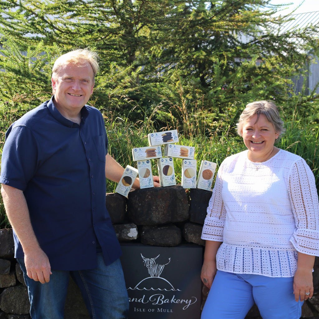 Couple standing outside next to stack of Island Bakery boxed goods on stone wall.
