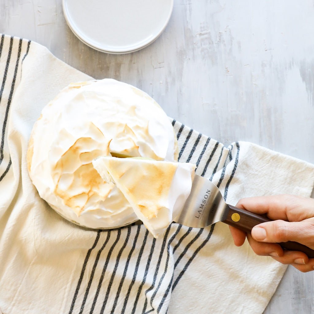 A hand holding a Lamson cake server with a wooden handle, lifting a slice of meringue-topped cake.