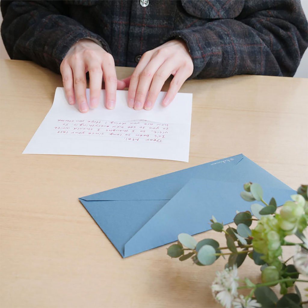 Person writing a letter with a blue envelope on a wooden table, accompanied by greenery.