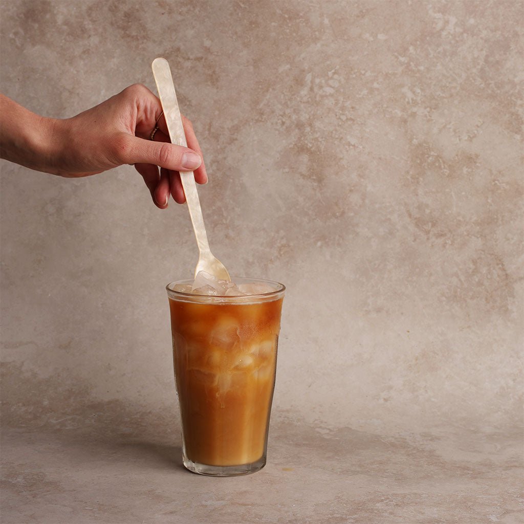 Hand stirring a glass of iced coffee with a cream acrylic spoon on a textured surface.