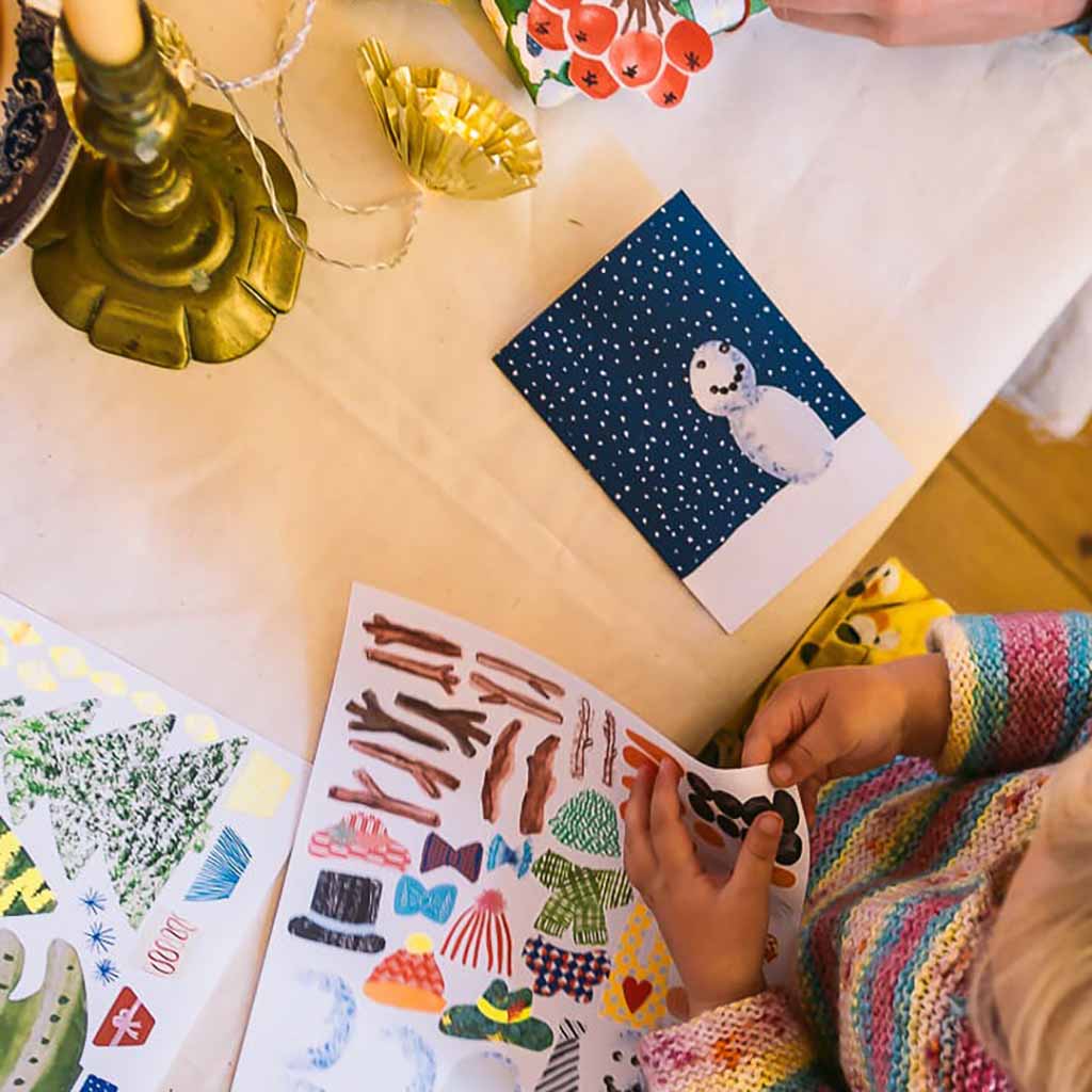 Child's hands holding a sticker over a sheet of stickers with a snowman card in the background.