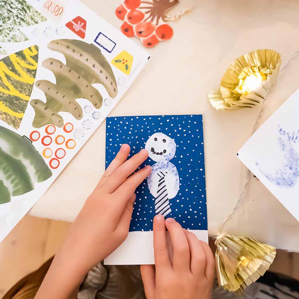 Child's hands holding a card with a snowman design on a table with craft materials.