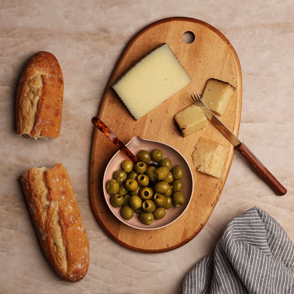 Wooden charcuterie board with cheese, bread and olives on a light background.