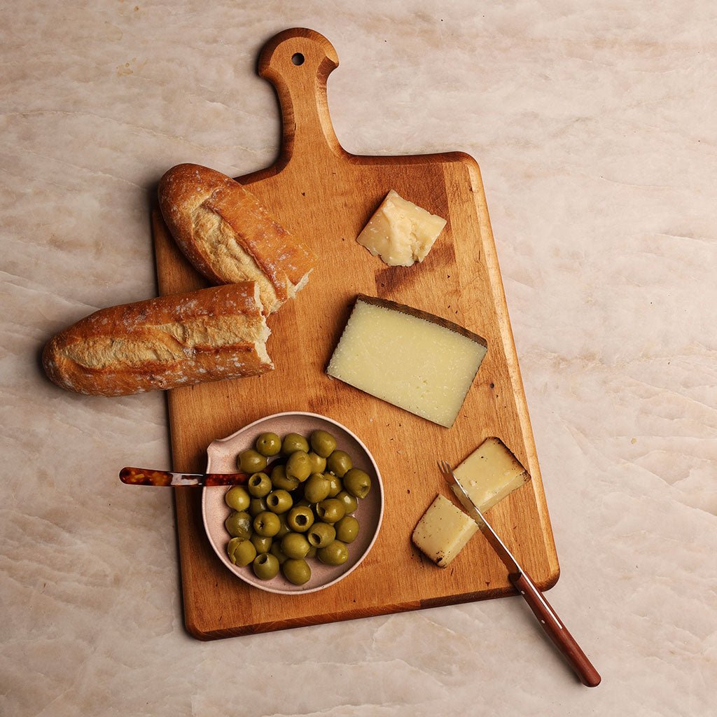 Wooden charcuterie board with cheese, bread and olives on a light background.