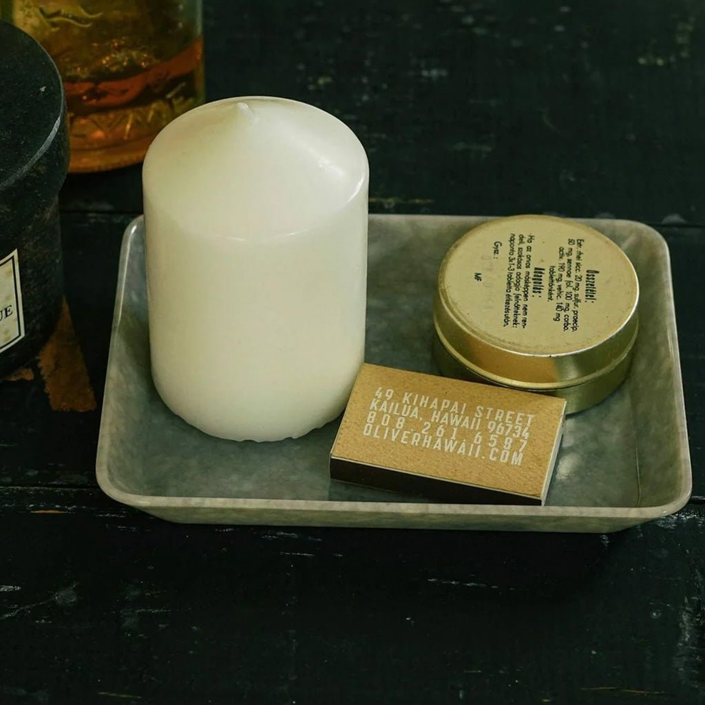 A white pillar candle displayed on a gray marbled tray alongside a small gold tin and a wooden matchbox on a dark tabletop.