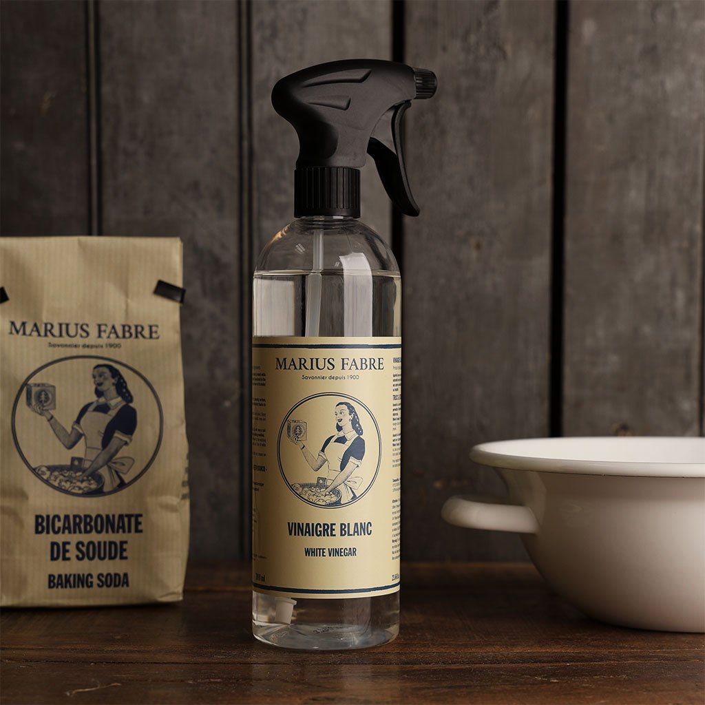 A clear spray bottle of Marius Fabre white vinegar stands on a wooden surface, with a paper bag of Marius Fabre baking soda to the left and a white enamel bowl to the right, all against a dark wood background.