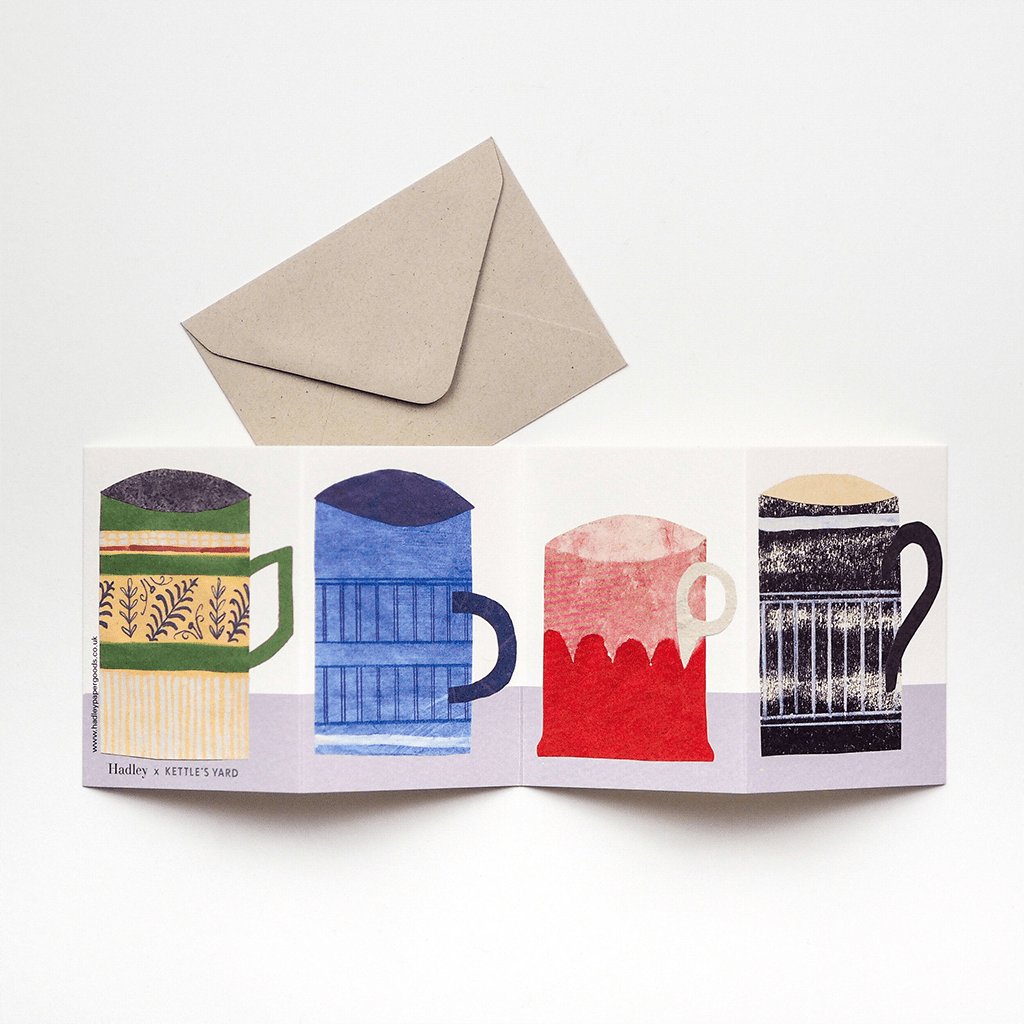 Accordion-style greeting card with four illustrated mugs in green, blue, red, and black, displayed open beneath a kraft paper envelope.
