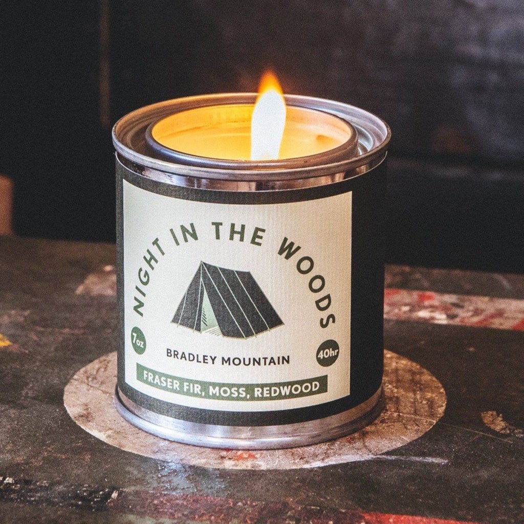Candle in a tin labeled 'Night in the Woods' by Bradley Mountain on a dark surface.