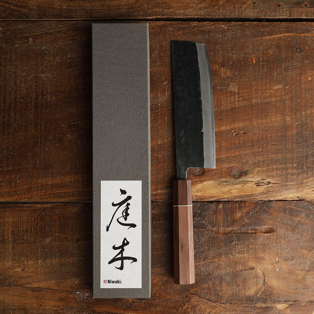 A Japanese-style kitchen knife with a dark steel blade and a brown wooden handle, displayed next to its textured gray box.