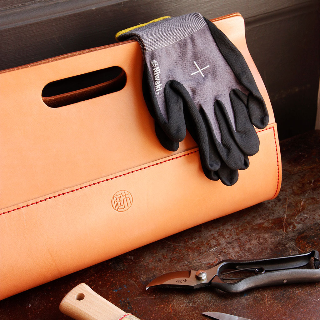 Niwaki garden gloves and tools next to leather tool bag on dark background