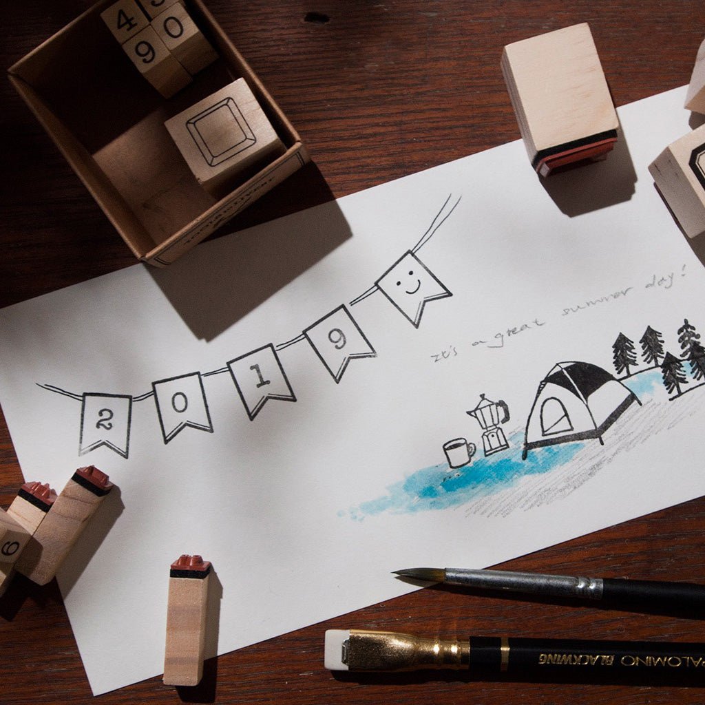A styled flat-lay of wooden rubber stamps and a paintbrush with a white sheet of paper featuring a hand-drawn 2019 summer camp scene and banner.