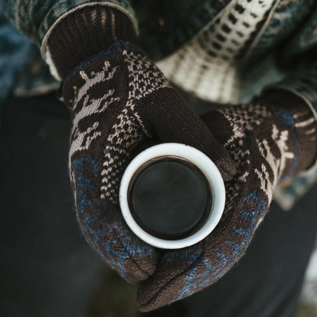 Hands wearing wool mittens and holding mug of coffee.