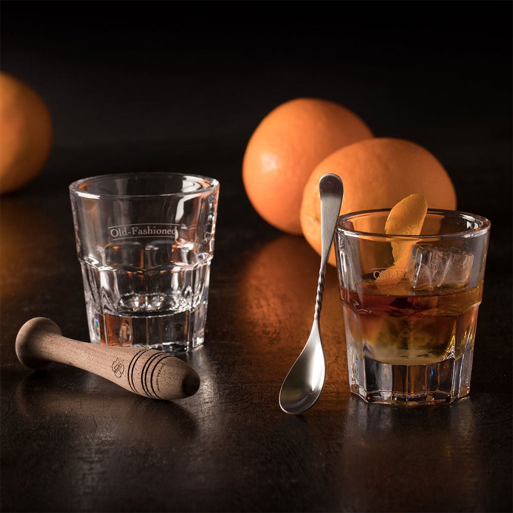 Two glasses with drinks, a spoon, and oranges on a dark surface.