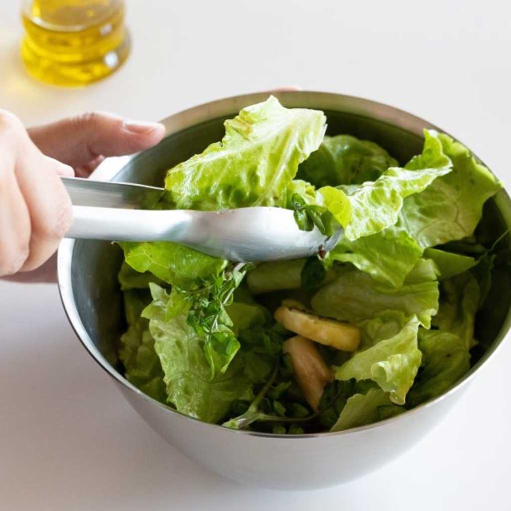 Person using tongs to serve a bowl of green salad on a white surface