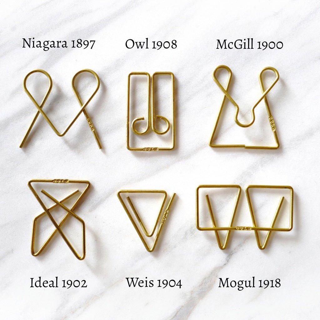 Six gold-colored metal wire paperclips arranged in a 2x3 grid on a white marble background, each labeled with its name and year: Niagara 1897, Owl 1908, McGill 1900, Ideal 1902, Weis 1904, and Mogul 1918.