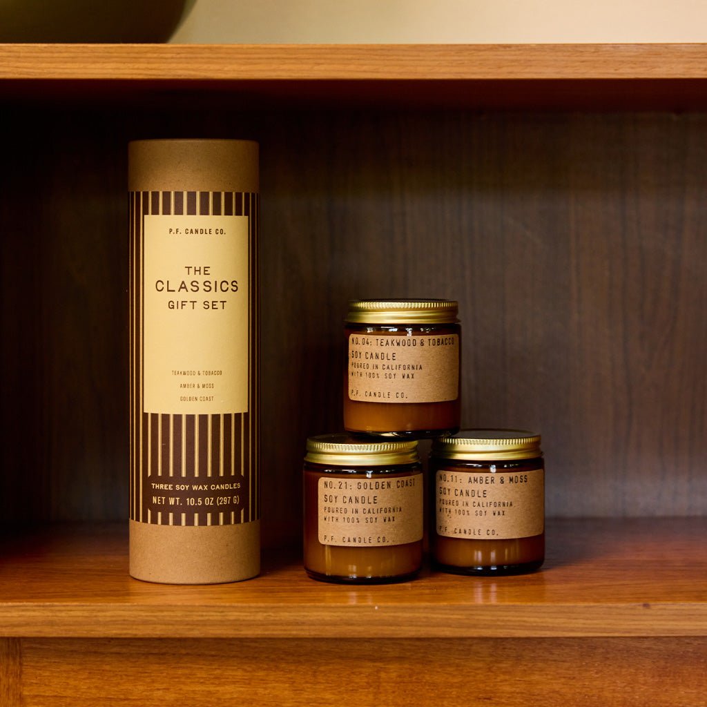 Three P.F. Candle Co. mini candles in amber jars with kraft labels and gold lids next to kraft tube packaging with festive green label on wooden shelf