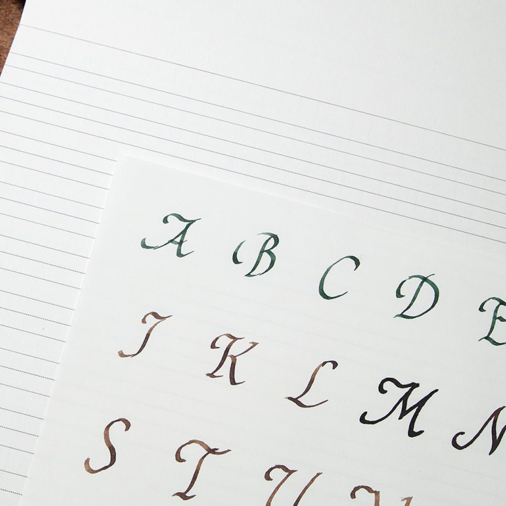 A close-up of a lined paper sheet showing a sample alphabet in elegant cursive script, with letters A through M visible in dark green, brown, and black ink.