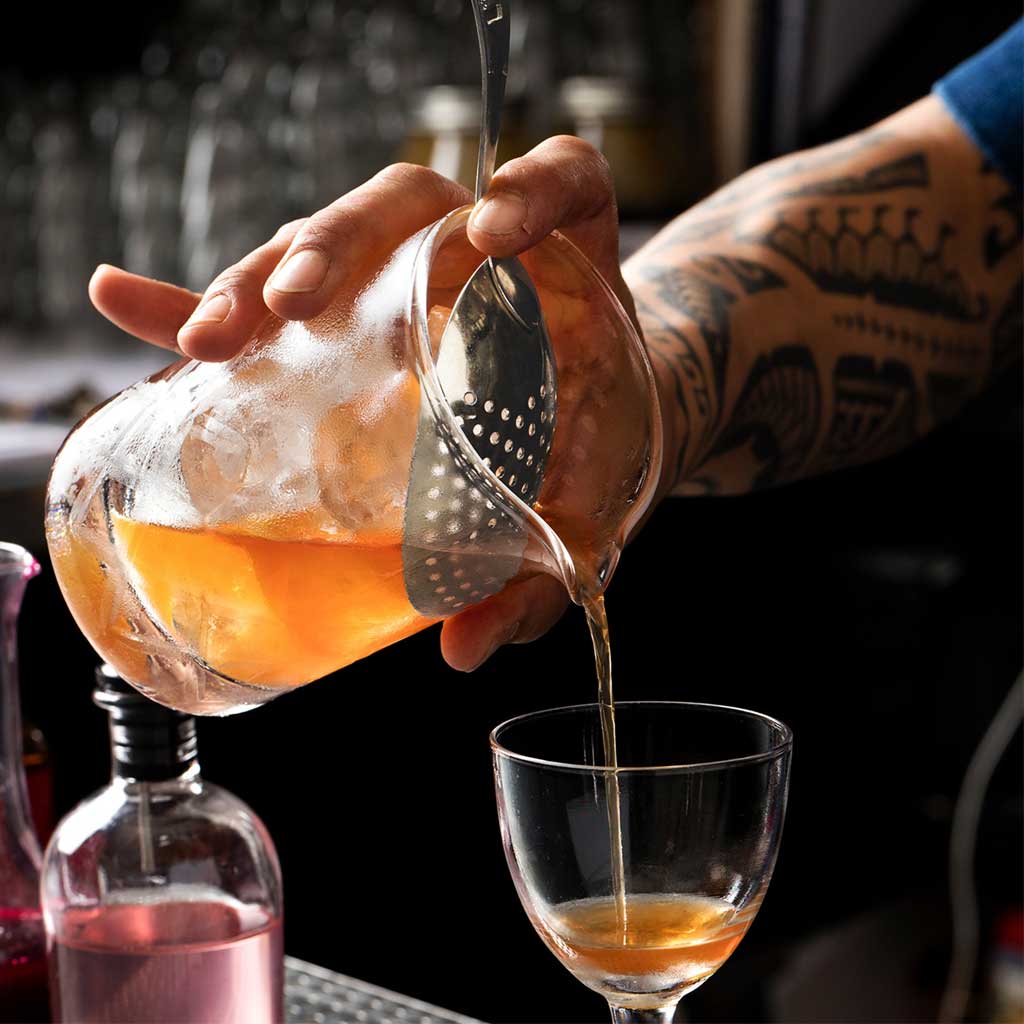 Person pouring a cocktail from a mixing glass through a julep strainer into a glass with a blurred bar setting in the background.