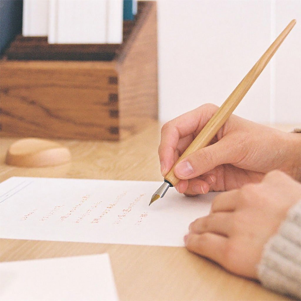 Hand writing with a fountain pen on paper, with a wooden desk and books in the background.