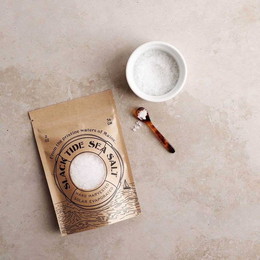 Kraft paper packaging of Slack Tide Sea Salt with a small bowl of salt and a spoon on a beige surface.